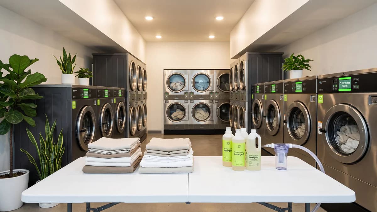 Clean modern laundromat with front-load washers, folded natural fabrics, biodegradable detergent bottles, microfiber filter, and green plants under bright lighting.